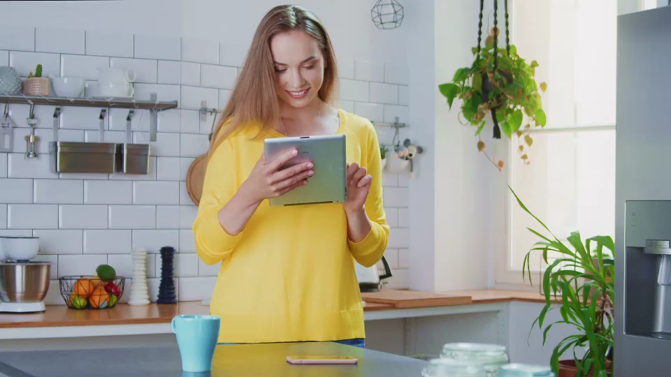 young woman in yellow sweater standing behind the table in the kitchen reading of the iPad, a still from online video advertisement