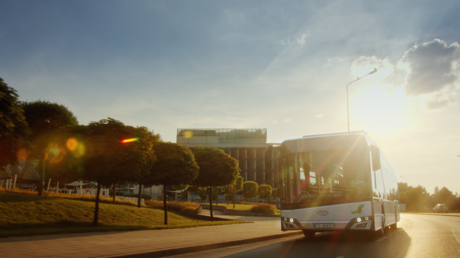 a shot from the product video showing a bus driving towards the camera with sunset behind it, a line of short trees by its side and modern building behind them